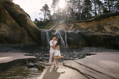Couple embracing on a sandy beach at a secluded coastal cove with a small waterfall cascading over rocky cliffs, pebbled shore, and sunlight filtering through trees.