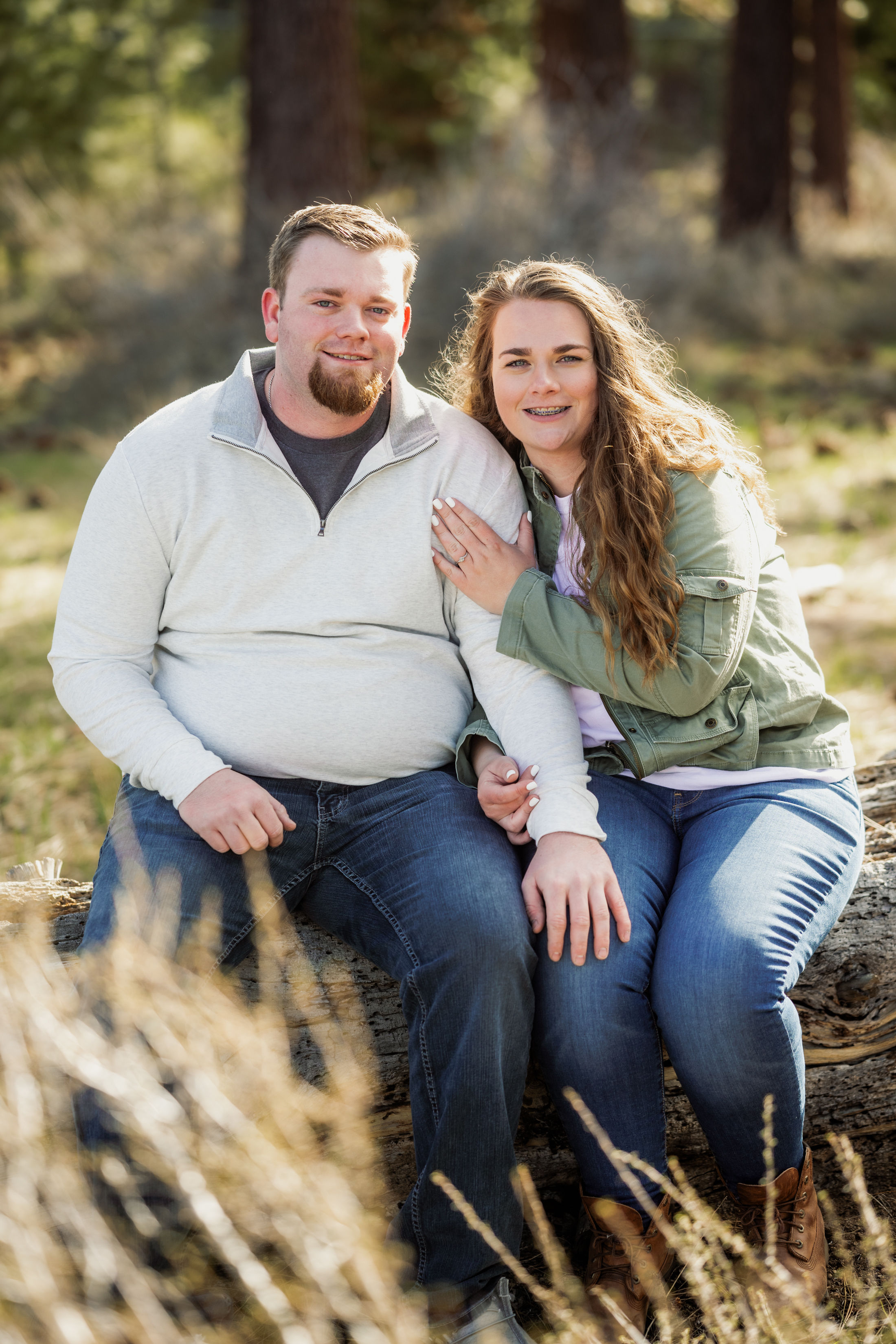 Smiling couple sitting on a fallen log in a sunlit pine-forest meadow, wearing casual jackets and jeans — cozy outdoor couple portrait