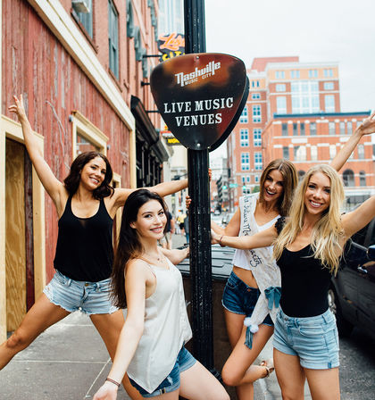 Four smiling women in summer outfits posing energetically around a 'Live Music Venues' sign on a downtown Nashville street