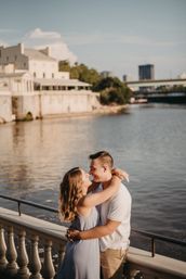 Romantic couple embracing on an urban riverfront promenade at golden hour, leaning on a balustrade with a historic riverside building and distant city skyline reflected in the water.