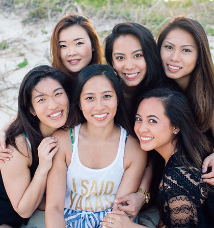 Six smiling women huddled together on a sandy beach in casual summer outfits, close-up group portrait with sand dunes in the background.
