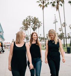 Three smiling women in casual black outfits walking a palm-lined California beach boardwalk, laughing beneath tall palm trees on an overcast day.