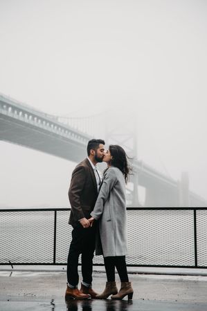 Romantic couple holding hands and kissing on a foggy urban waterfront promenade with a suspension bridge looming in the mist