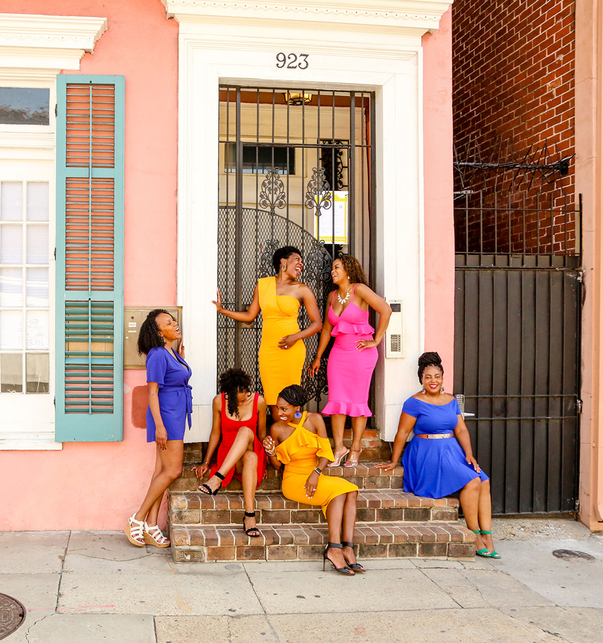 Six women in vibrant summer dresses laughing and posing on brick steps outside a pastel pink city townhouse with teal shutters and a decorative iron door — colorful group portrait on urban street