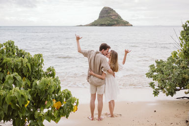 Couple kissing on a sandy tropical beach, framed by green foliage, both making shaka hand gestures while facing a small lush island across the calm ocean.
