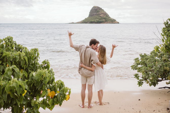 Couple kissing on a sandy tropical beach, framed by green foliage, both making shaka hand gestures while facing a small lush island across the calm ocean.
