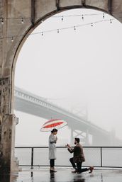 Man kneeling to propose under a red-and-white umbrella at a foggy waterfront by a large suspension bridge, framed by a weathered stone arch and string lights.