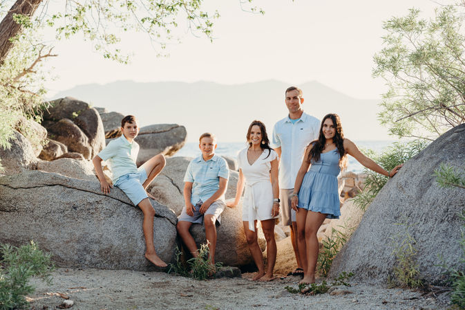 Sun-kissed family portrait of five in casual summer outfits on a rocky mountain-lake shoreline at golden hour, posed on large boulders with sparse trees and distant mountains