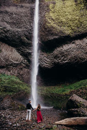 Couple holding hands at the base of a tall, slender waterfall cascading down textured basalt columns into a mossy, rocky canyon, woman in a flowing red dress.