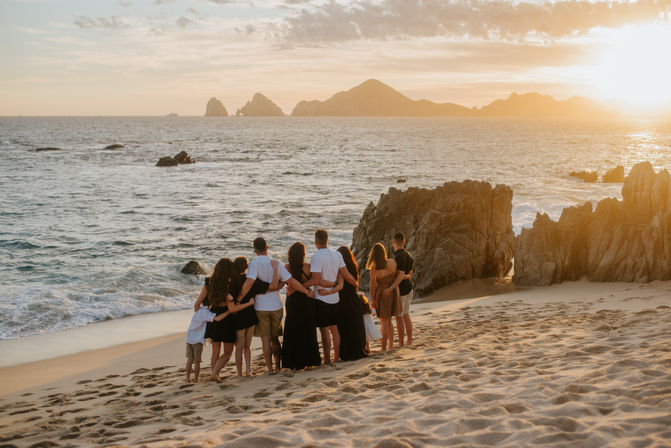 Sun-kissed family group embracing on a sandy beach at sunset, silhouetted against ocean waves and rocky islands on the horizon.