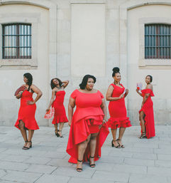 Five women in red cocktail dresses and heels striking confident poses with handheld fans on a paved city plaza in front of a pale stone building with arched windows.