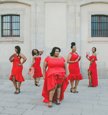 Five women in red cocktail dresses and heels striking confident poses with handheld fans on a paved city plaza in front of a pale stone building with arched windows.