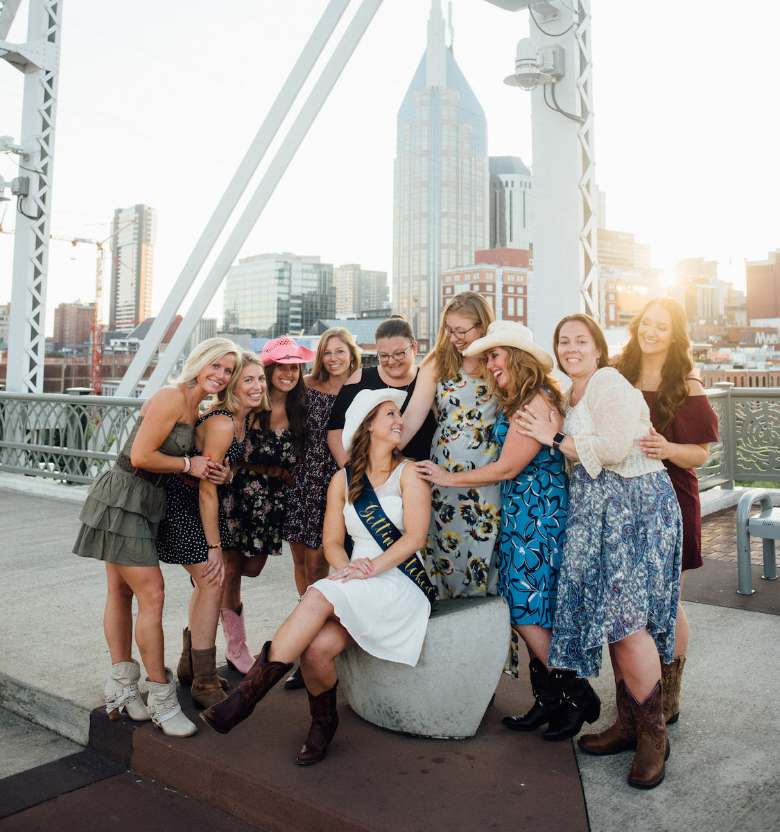 Bachelorette party on a downtown bridge at sunset — bride-to-be in a white dress and sash seated while friends in floral dresses, cowboy boots and hats laugh together with the city skyline behind them.