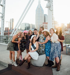 Bachelorette party on a downtown bridge at sunset — bride-to-be in a white dress and sash seated while friends in floral dresses, cowboy boots and hats laugh together with the city skyline behind them.