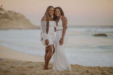 Two smiling women in white dresses embracing on a sandy beach at sunset, with gentle ocean waves and cliffs in the soft background.
