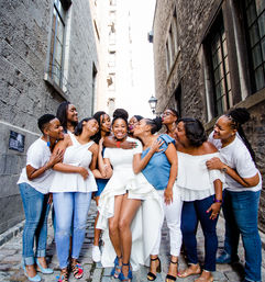 Smiling bride in a high-low white dress embraced and kissed by laughing friends on a narrow cobblestone alley flanked by tall historic stone buildings