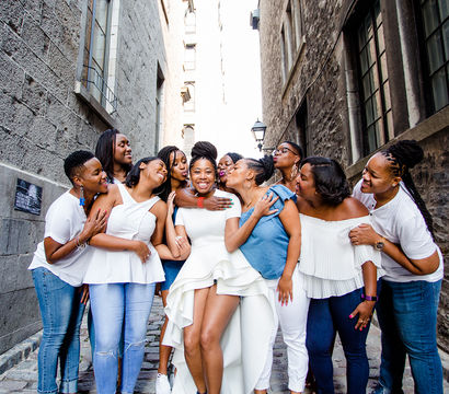 Smiling bride in a high-low white dress embraced and kissed by laughing friends on a narrow cobblestone alley flanked by tall historic stone buildings