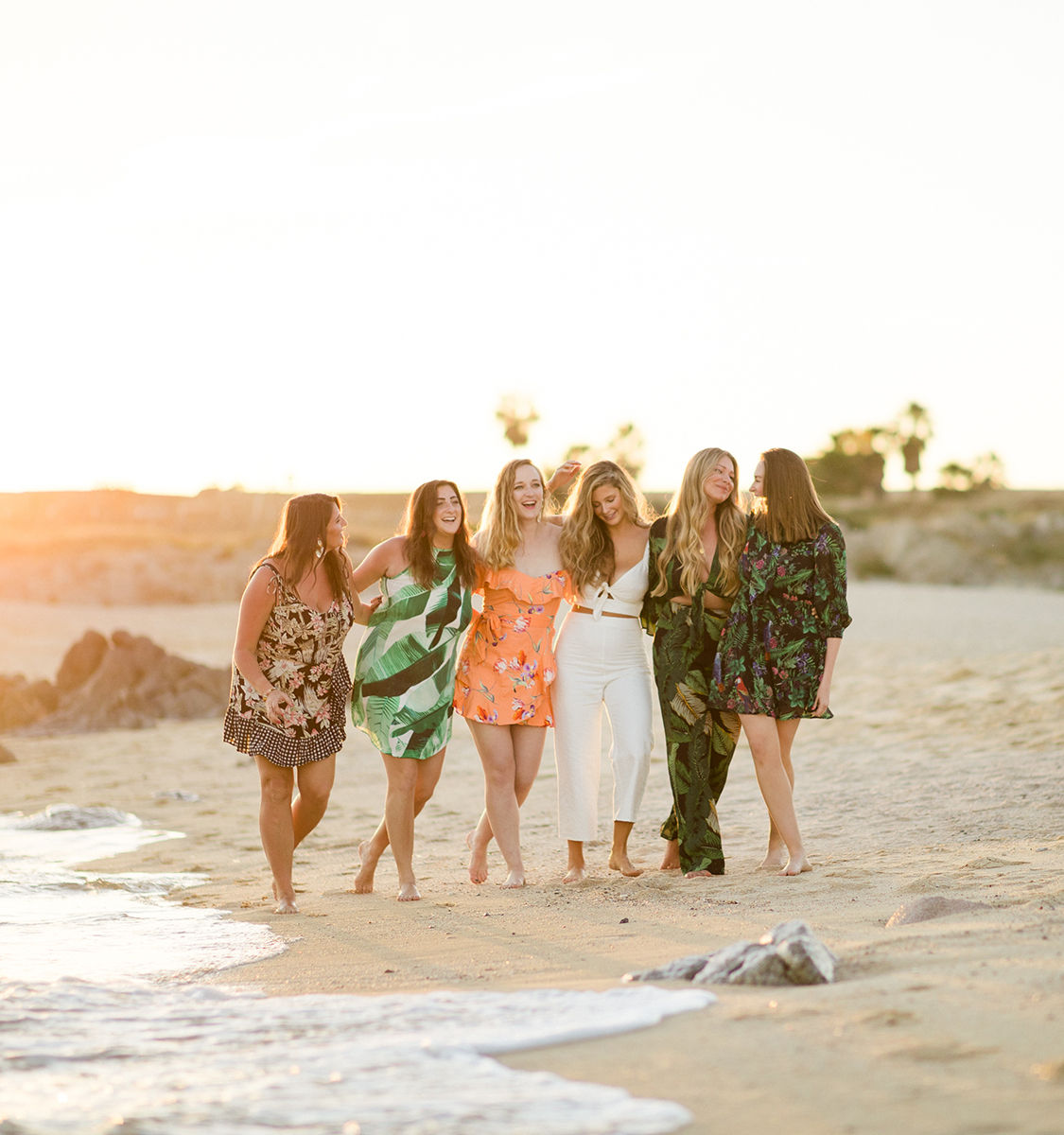 Six friends walking barefoot along a sunlit beach at sunset, laughing and linking arms in colorful summer outfits with palm trees in the background.