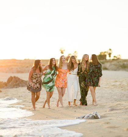 Six friends walking barefoot along a sunlit beach at sunset, laughing and linking arms in colorful summer outfits with palm trees in the background.
