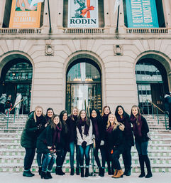 Group of smiling young women in winter coats and matching maroon scarves posing on snowy steps outside a grand city museum entrance with arched doorways and colorful exhibition banners.