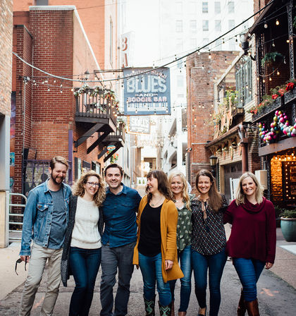 Seven friends walking arm-in-arm down a lively downtown brick alley in a historic entertainment district, smiling under string lights and colorful storefronts with a vintage music bar sign.