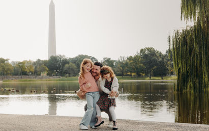 Smiling father hugging two young daughters by a pond with the Washington Monument and willow trees in the background, Washington, D.C.