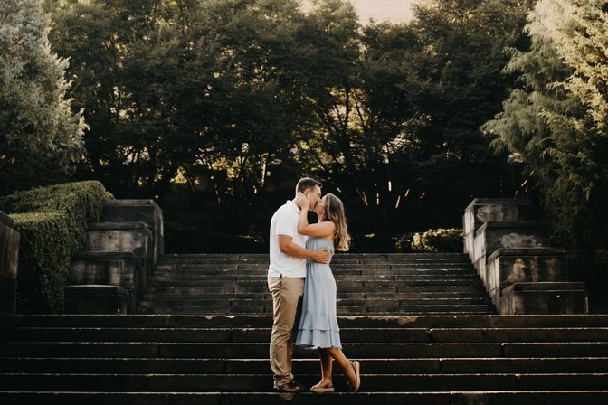 Romantic couple embracing and kissing on wide stone steps in a leafy park at golden hour, framed by trees and formal stone terraces.