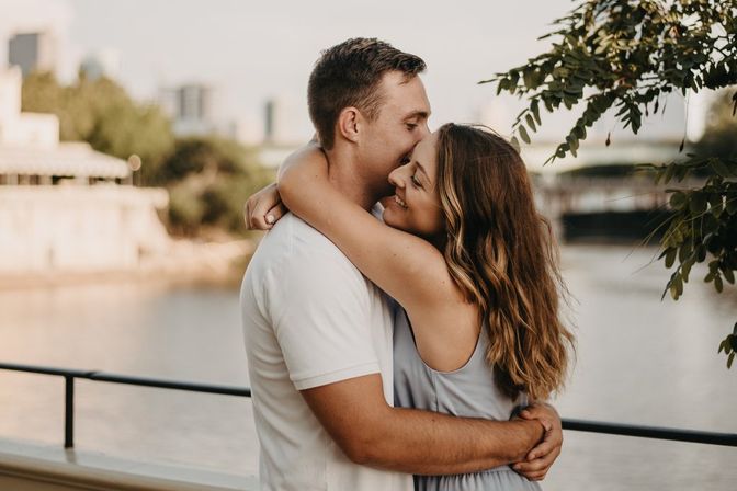 Smiling young couple hugging on an urban riverfront railing at golden hour, woman with wavy hair embracing a man in a white T‑shirt with bridge and city skyline softly blurred in the background.