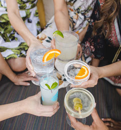 Top-down view of a group toasting colorful cocktails and mocktails with orange, lime, mint and olive garnishes on a striped outdoor patio table, summer brunch vibe.