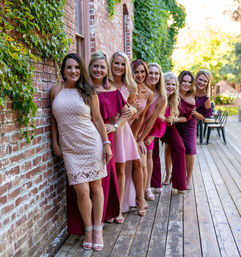 Eight women in pink and burgundy dresses smiling and leaning in a line along an ivy-covered brick wall on a sunlit wooden deck — playful outdoor group portrait.