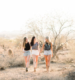 Three young women walking hand-in-hand on a sunlit Southwestern desert trail at golden hour, wearing casual tops, denim shorts and boots amid dry shrubs and cacti.