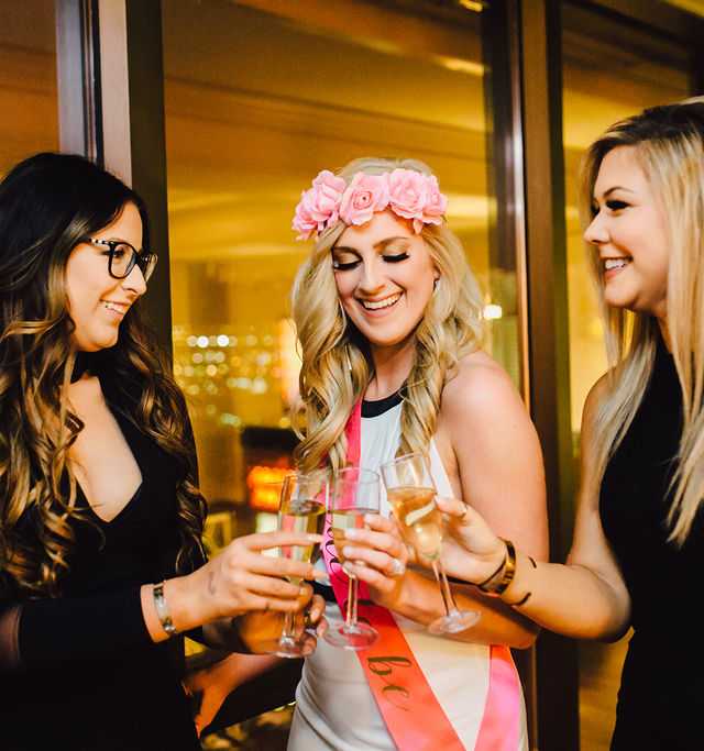 Bachelorette party scene: smiling bride-to-be in a pink flower crown and sash toasting champagne with two friends by a city-lit hotel window