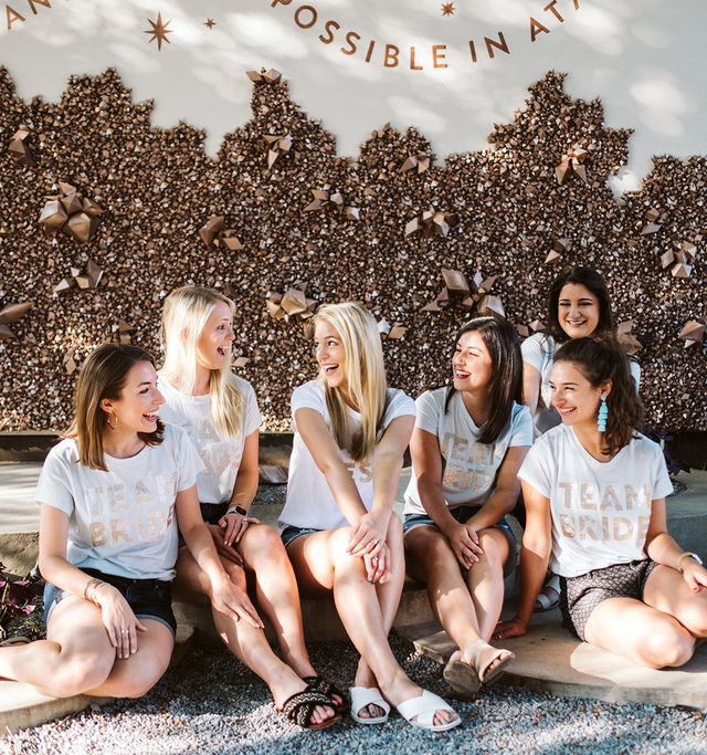 Sunlit bachelorette group of six women in matching white "Team Bride" tees laughing on a stone bench against a metallic starburst mural backdrop.