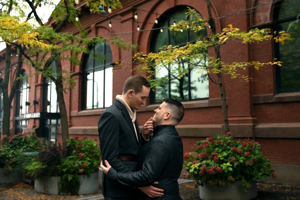 Two men sharing a tender embrace on an urban sidewalk in front of a red-brick building with arched windows, string lights overhead and potted autumn flowers.