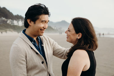 Smiling couple facing each other on a misty sandy beach with coastal houses and hills in the background — romantic seaside portrait.