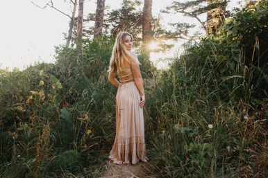 Woman in a flowing boho beige dress on a grassy woodland path, looking back as golden-hour sun filters through trees.