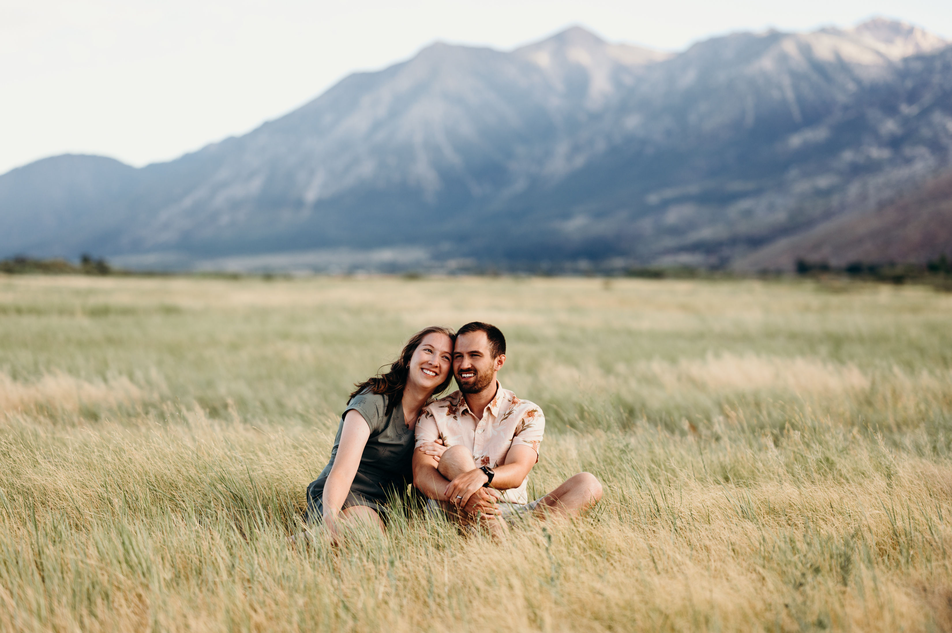 Smiling couple seated in tall golden grass of a mountain meadow, leaning together with rugged peaks and open sky in the background