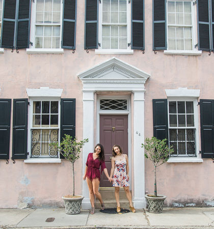 Two friends holding hands and smiling in front of a historic pastel-pink townhouse with black shutters, white trim, a mauve door, and potted trees.