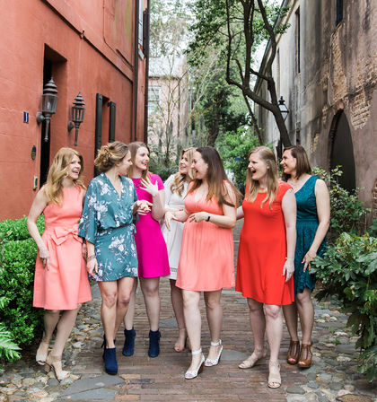 Seven women in colorful dresses laughing and walking together on a cobblestone alley lined with brick buildings and greenery in a tree-lined historic district