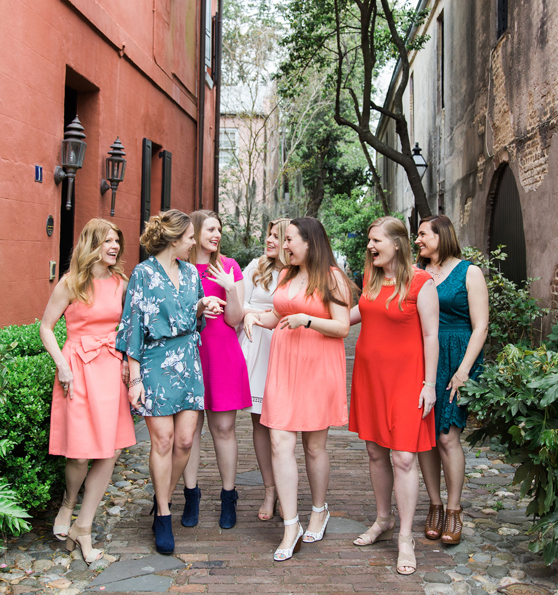 Seven women in colorful dresses laughing and walking together on a cobblestone alley lined with brick buildings and greenery in a tree-lined historic district