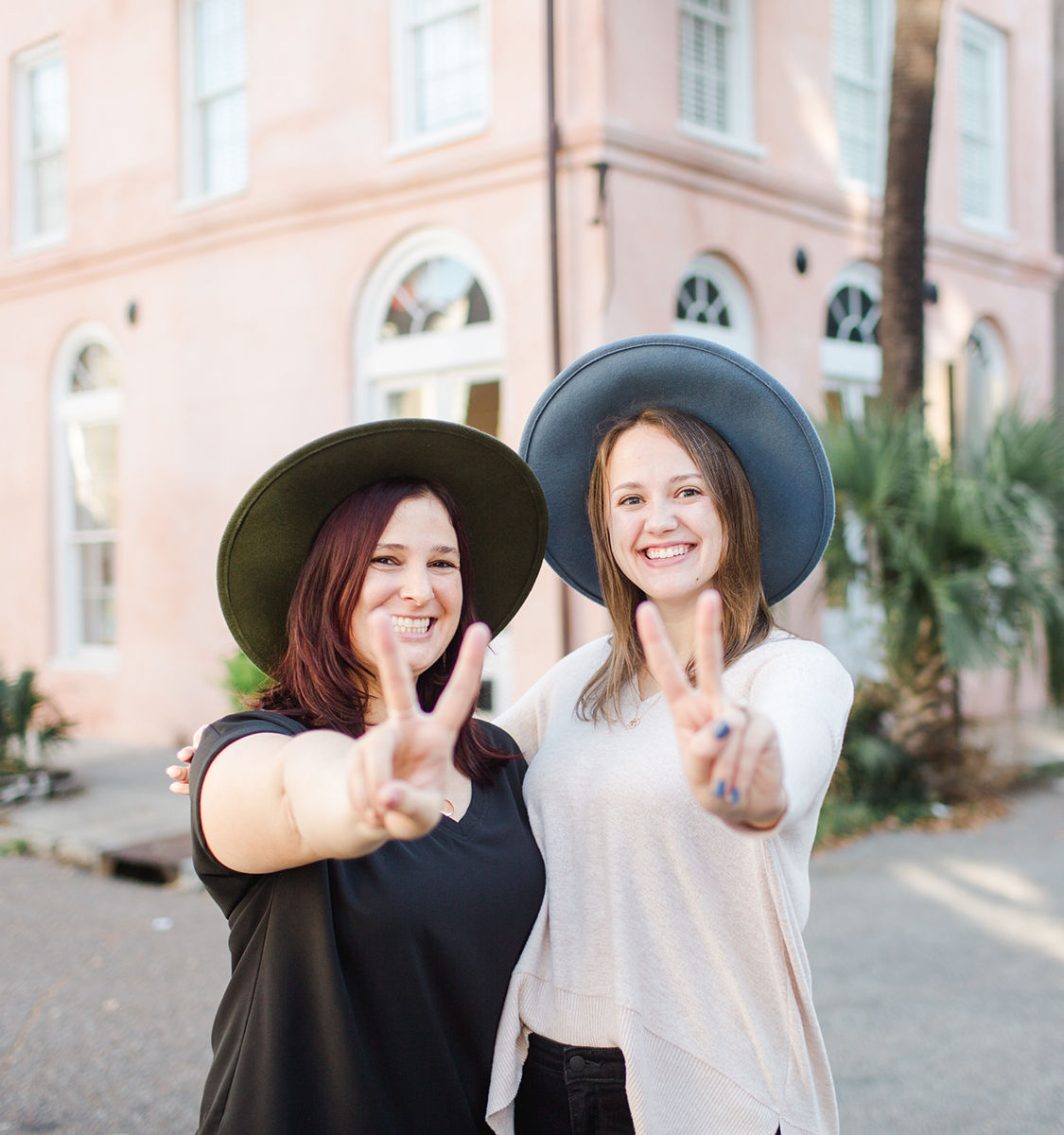 Two smiling women wearing wide-brim hats flashing peace signs in front of a pastel pink building with arched windows and palm trees