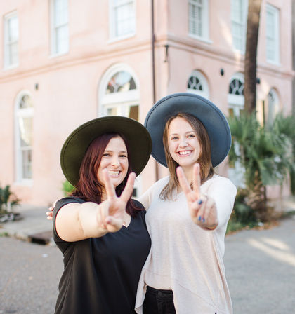 Two smiling women wearing wide-brim hats flashing peace signs in front of a pastel pink building with arched windows and palm trees