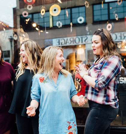 Three women laughing and chatting outside a downtown shop window with colorful hanging disc decorations, a candid urban shopping scene.