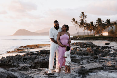 Pregnant woman in a pink dress with her partner and young child embracing on a rocky tropical beach at sunset, palm trees and distant mountains in the background — family maternity portrait.