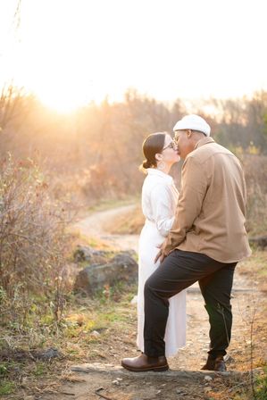 Couple sharing a kiss on a sunlit woodland trail at golden hour, warm autumn light and a winding path in the background.