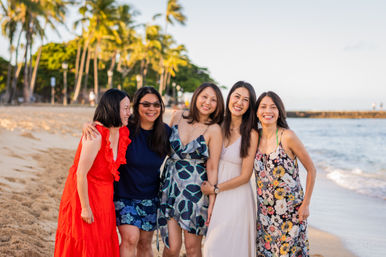 Five smiling women in colorful summer dresses posing together on a palm‑lined tropical beach by the ocean at sunset.