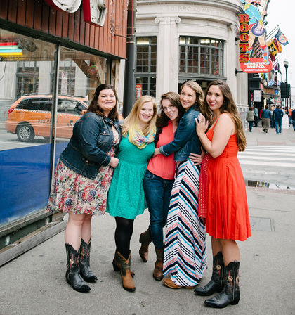Five friends in colorful dresses, denim jackets and cowboy boots posing on a downtown entertainment-district sidewalk in front of a historic theater and storefronts