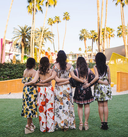 Five friends standing arm-in-arm on a sunny palm-tree-lined resort lawn, wearing colorful floral summer dresses with retro brightly painted hotel facades in the background.