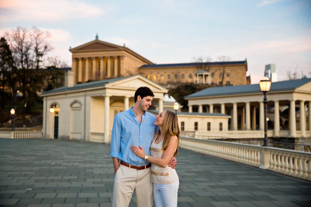 Young couple embracing on a wide stone promenade at dusk with a neoclassical museum and colonnaded buildings in the background, decorative lampposts and a soft evening sky.