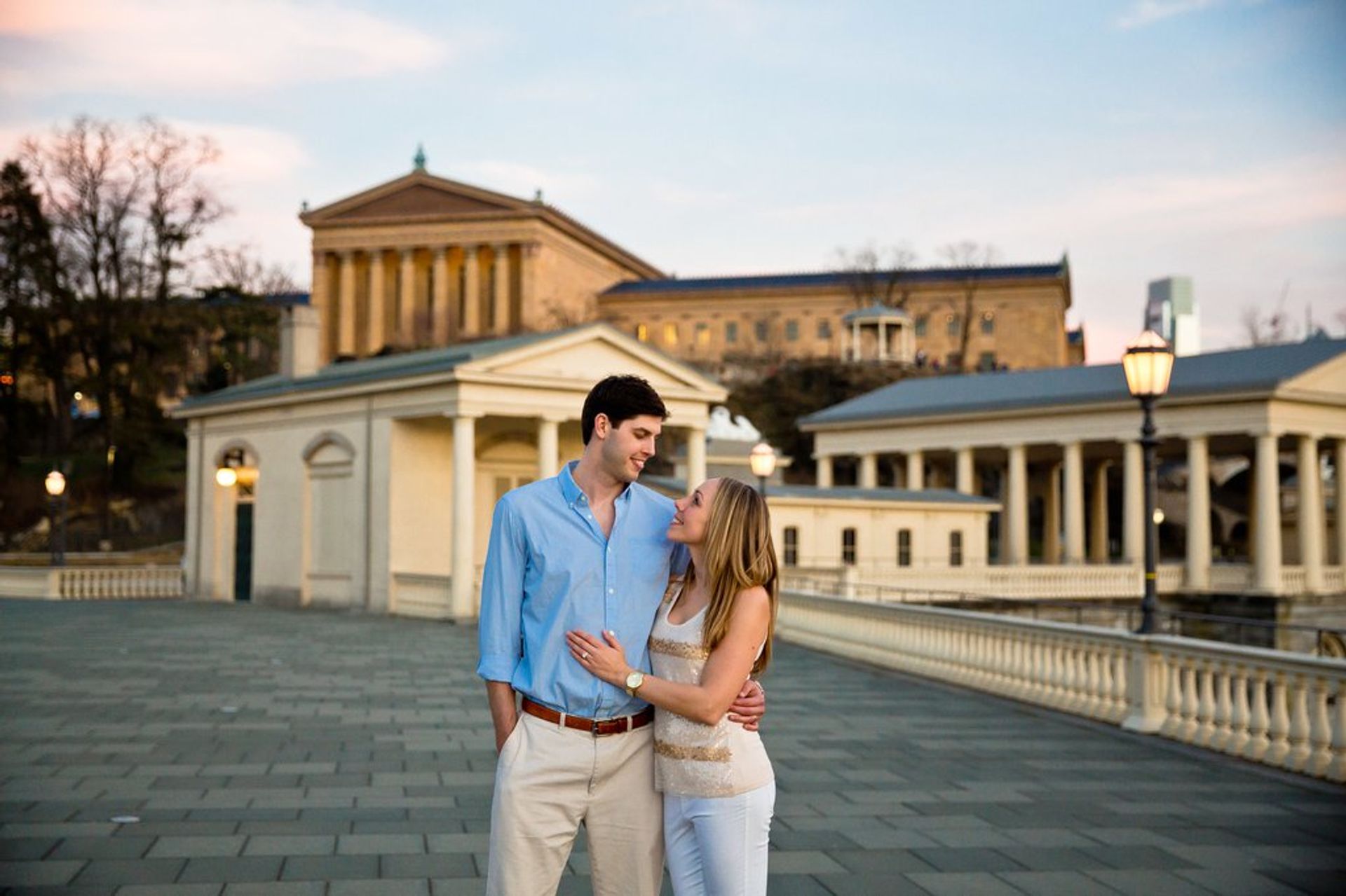 Young couple embracing on a wide stone promenade at dusk with a neoclassical museum and colonnaded buildings in the background, decorative lampposts and a soft evening sky.